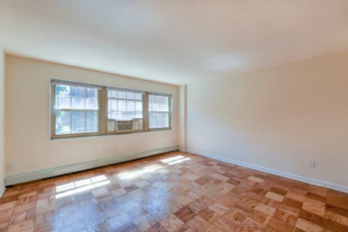 An empty room with parquet flooring, large windows, and an air conditioning unit installed. Walls are painted beige. | DC Apartments by WC Smith