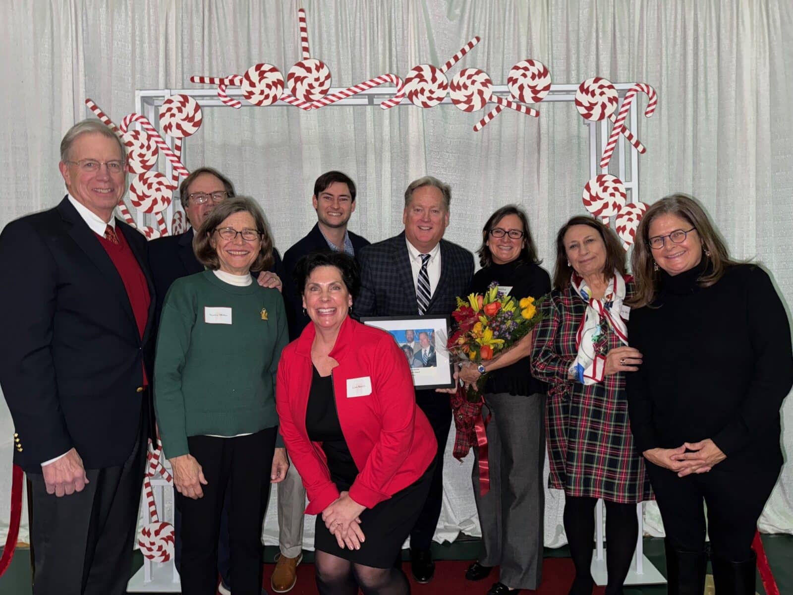 A group of people poses for a photo in front of a white curtain adorned with candy cane decorations. One holds a framed picture of the Bill Smith Award, while another clutches a bouquet of flowers. Pat McKenzie stands proudly among them, capturing the spirit of celebration. | DC Apartments by WC Smith