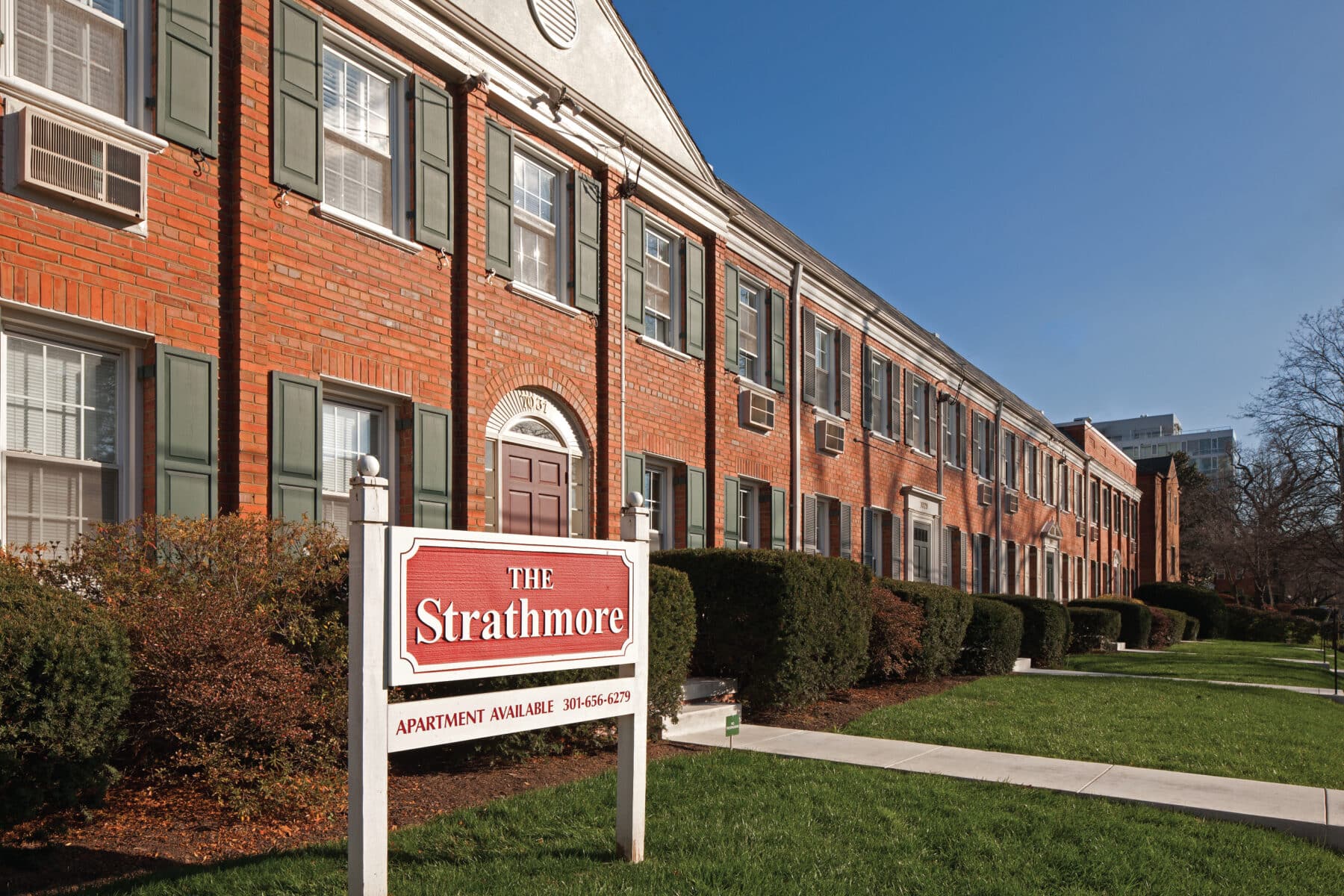 Red-brick apartment building with green shutters, labeled "The Strathmore." A sign in front includes contact information. A sunny day with clear blue sky. | DC Apartments by WC Smith