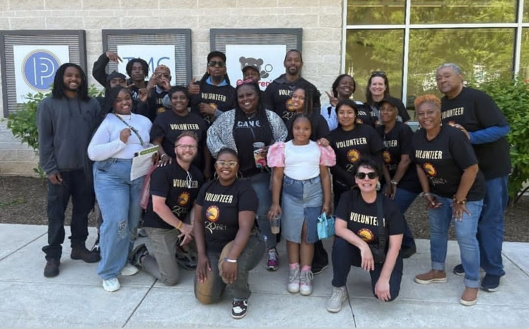 A group of adults and children, many wearing "Volunteer" shirts, pose and smile together outdoors in front of a building during THEARC's 20th Anniversary Spring Fling. | DC Apartments by WC Smith
