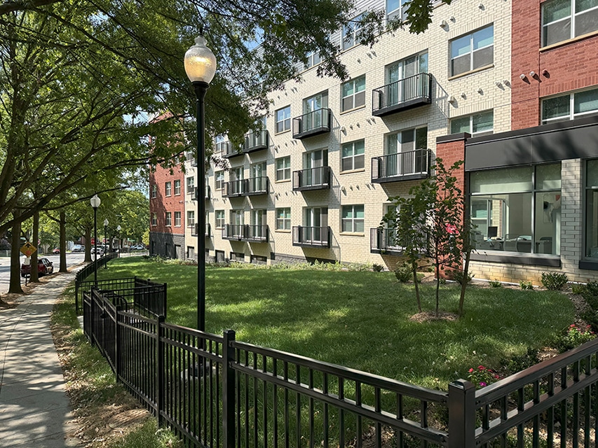 A modern WC Smith apartment building with small balconies overlooks a grassy lawn, black metal fence, and tree-lined sidewalk on a sunny day, reflecting its commitment to quality living and potential LEED Certification standards. | DC Apartments by WC Smith