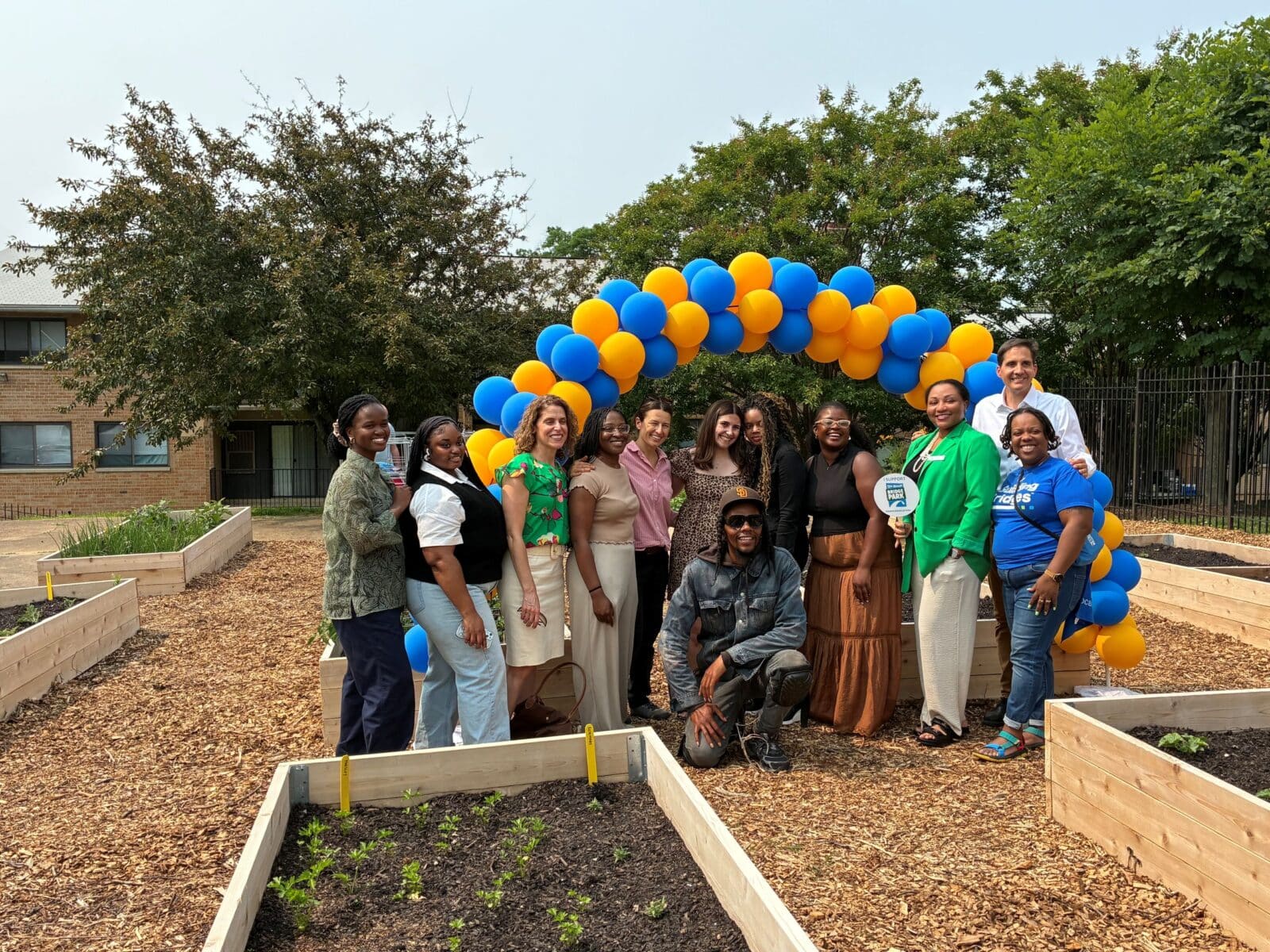 A group of people pose and smile in front of a blue and yellow balloon arch at the 11th Street Bridge Park community garden, surrounded by raised beds and wood chip pathways. | DC Apartments by WC Smith