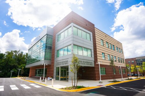 A modern three-story building with glass and brick exterior, labeled "The Washington School for Girls," under a partly cloudy sky. | DC Apartments by WC Smith