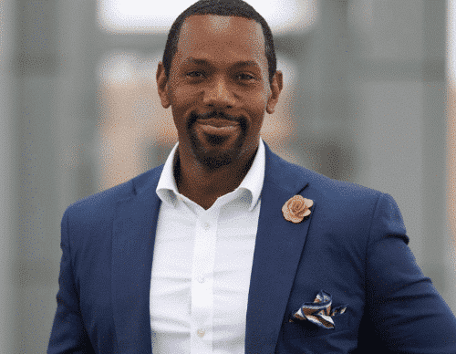 Rahsaan Bernard, Chief Operating Officer at WC Smith, stands outdoors in a blue suit and white shirt, wearing a beige flower lapel pin and patterned pocket square, smiling warmly at the camera. | DC Apartments by WC Smith