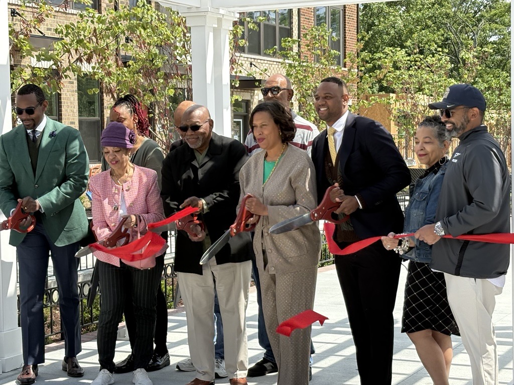 Eight people, including Community Leader Karen Williams, stand in a row cutting a red ribbon with oversized scissors at Skyland Town Center Park's outdoor ribbon-cutting ceremony. Trees and brick buildings are in the background. | DC Apartments by WC Smith